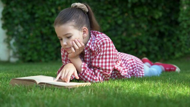 Happy little girl, in a red shirt, lying on a green carpet, lawn. smiling child reads a book lying on the grass. the concept of a carefree childhood, long-awaited summer, summer holidays.