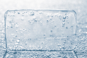 The rectangular block of pure transparent ice with air bubbles in cold blue tones on a winter river ice, covered with white snow.