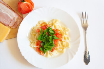 Pasta farfalle with tomatoes and herbs in a plate, fork, bacon, cheese and tomato on a white background, top view