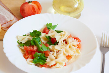 Pasta farfalle with tomatoes and herbs in a plate, fork, bacon, cheese, tomato and oil in a jug on a white background