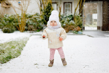 Happy little baby girl making first steps outdoors in winter through snow. Cute toddler learning walking. Child having fun on cold snowy day. Baby's first snow, activity. Winter walk outdoors