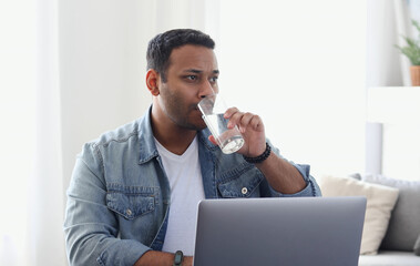 Focused indian businessman working using laptop, resting and drinking refreshing water for more effective work, remote work at home