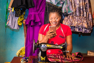 Fototapeta na wymiar African female tailor at work with colorful clothes using mobile phone