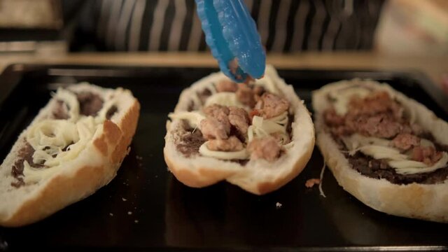 Blue tongs adding chopped meat to traditional Mexican molletes above black tray