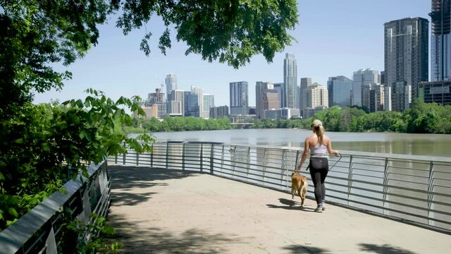 A woman in a hat and exercise clothing walks her dog on a sunny day down the lakeside boardwalk in Austin, Texas with skyline in the background.