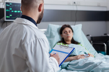 Specialist physician with stethoscope writing disease treatment on clipboard checking sickness symptom while working in hospital ward. Sick woman patient resting in bed discussing illness recovery