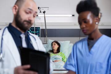 Practitioner doctor with black nurse checking recovery treatment using tablet computer working in hospital ward. Hospitalized patient woman resting in bed discussing with specialist physician