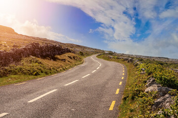 Small narrow asphalt road in Burren area of Ireland. Warm sunny day. Travel and tourism concept. Nobody. Clear cloudy sky. Sun flare