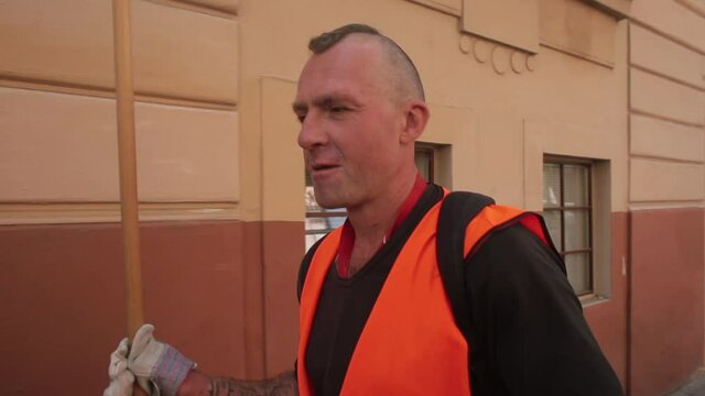 A man in an orange vest and gloves speaks on the move on a city street. The cleaner carries a tool in his hand. Camera movement.