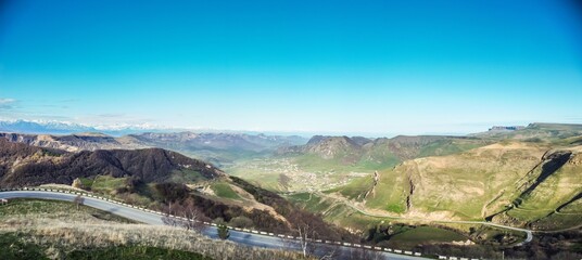 Naklejka premium Aerial panoramic landscape view over Gum-bashi mountain pass, with green grass on cliffs, winding road on hills, Сaucasus mountian range and Elbrus mountain. Karachay-Cherkessia