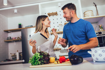 Young happy couple is enjoying and preparing healthy meal in their kitchen and reading recipes on the digital tablet.