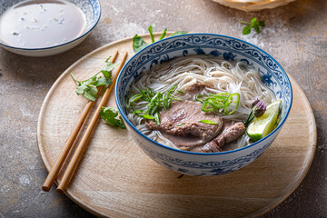 Vietnamese noodle soup pho bo in bowl, garnished with basil, mint, lime, on concrete background - obrazy, fototapety, plakaty