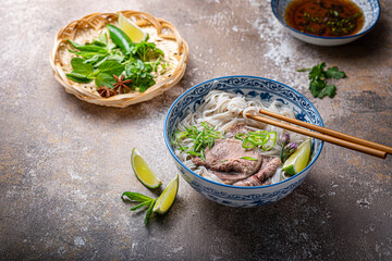 Traditional vietnamese noodle soup pho in bowl, garnished with basil, mint, lime, on concrete background - obrazy, fototapety, plakaty