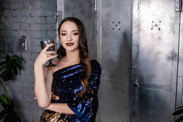 Close-up portrait of a beautiful girl with an evening festive, professional make-up, holding a glass of wine. Young woman in a blue and gold dress in sequins on a metallic, gray shiny background