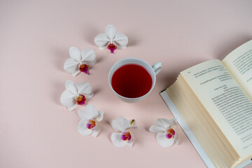 A cup of herbal tea with orchid flowers and a book on a pink background