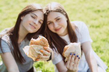Two young women friends eating delicious burgers in the park on the grass. Not a healthy diet. Soft...