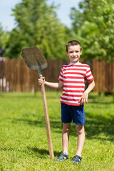 A little cheerful boy stands and holds a shovel in his hand, ready to work in the garden of a country house.