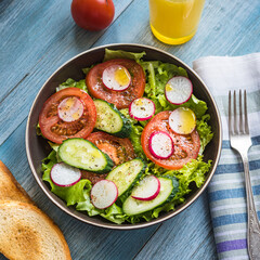 Vegan breakfast - salad with tomatoes, cucumbers and radishes dressed with ground pepper and olive oil in a plate and a glass of orange juice on the table, top view