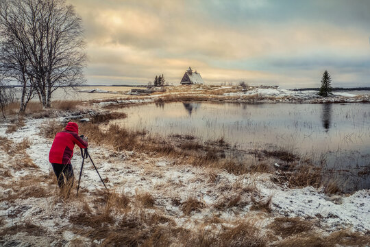 Photographer shoots a wonderful dawn snowy winter landscape with authentic house on the shore.