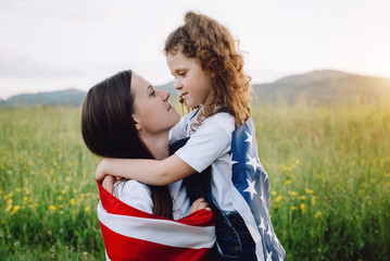 Close up of caring young mother and cute small daughter kid sit in grass with national US flag outdoors on background green mountains during sunset. American flag, country, patriotism, july fourth