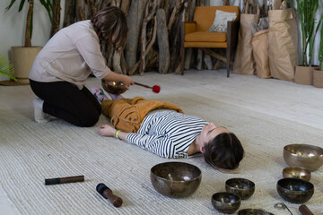 Singing bowls massage at home: two women practice tibet sound therapy together using vibrations and music from traditional bronze bowls and gong of tibetan alternative medicine for meditation concept
