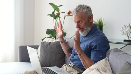 Modern middle-aged bearded man using laptop computer for virtual meeting sitting on the couch at home. Mature grey-haired man waving into webcam, chatting online, holding video call