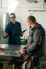 A tinsmith is measuring in his workshop with a colleague engineer who is taking notes on tablet