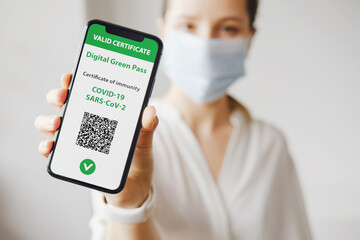 Young caucasian woman in mask holding smartphone with digital green pass and QR code on the screen....