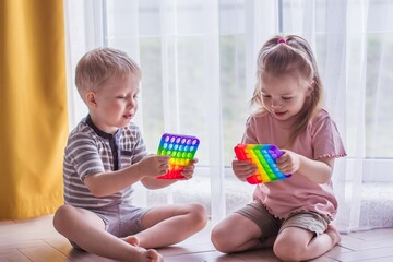 Blonde boy and girl children sit on the floor near the window and play silicone toy antistress pop it. Pop it sensory toy. Stress relief. Colorful anti stress silicone sensors toy - obrazy, fototapety, plakaty