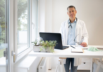 handsome doctor with white coat and blue shirt and stethoscope stands behind high table and works on laptop and has web conference or meeting