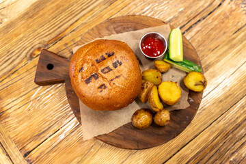 Fototapeta premium Burger with meat served with cucumbers, cetchup and fried baby potato on round cutting board. Unhealty dinner