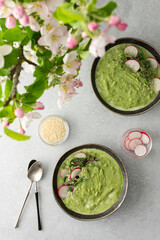 Top view of a table with two plates of creamy spinach broccoli soup, soup decorated with microgreens and sesame seeds with radishes, a bouquet of flowering apple tree branches