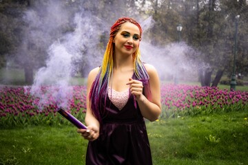 A girl with a smile with bright makeup and colored braids in a purple dress blows artificial purple smoke in a spring blooming park