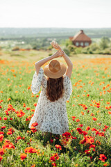 Girl wearing white dress and straw hat walks among red poppy flowers on the sunset.