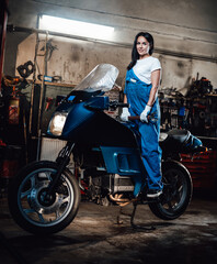 Fototapeta na wymiar Young female mechanic in blue overalls posing on sportbike in garage or workshop