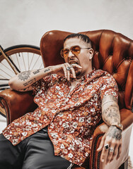 Tattooed gangster yawns while sitting on a luxurious retro armchair, looks very expressive and freakly