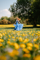 A happy fairy-tale girl in an airy blue dress runs joyfully along the green lawn in the park. Blooming field in yellow dandelions. Allergy free concept.