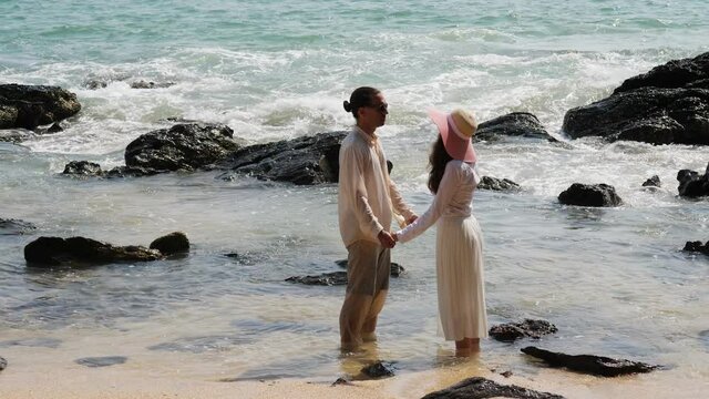 Couple at beach, stand in water and hold hands, look to each other. Wave break on rough stones and rush up, people stay still, not take eyes off. Tourist pair at vacation time in Thailand