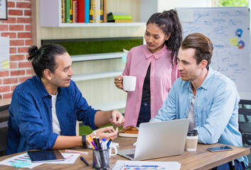 Three multiracial diverse cheerful Caucasian and Asian business people comfortably drinking coffee, smiling, talking together with happiness and relaxation in the morning at cozy modern office.