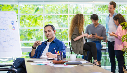 Foreground focus handsome hipster Caucasian male creative director, boss or leader sitting in comfortable modern office, smiling with confidence and profession with background of mixed race employee.