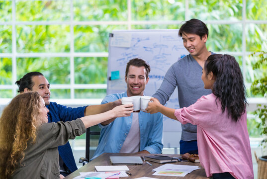 Multiethnic diverse happy casual businesspeople smiling with happiness and comfort, clinking cups of coffee together to congratulate successful creative marketing project, standing in cozy office.