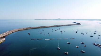 Sunny Holyhead harbour breakwater landmark maritime yachts docked along transparent calm blue shoreline aerial view dolly right