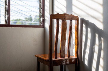 Behind the back of a wooden chair with the sunlight louvered windows onto the wall of the room.