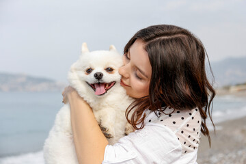 Young woman hugging her pomeranian spitz on the beach. Female bonding with white coated pom dog outside on the walk. Close up, copy space for text, ocean background.