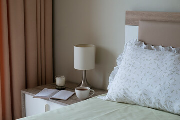 Close up shot of the nightstand with a book and coffee cup on top near the unmade bed. Good morning concept. Bedroom full of natural light. Copy space for text, background, top view.