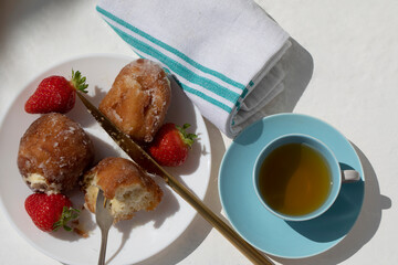 SUNLIT SUGAR DONUTS STRAWBERRIES SWEET TREAT JASMIN TEA BLUE VINTAGE CUP & SAUCER GOLD KIFE SMALL FORK WHITE CLOTH ON WHITE SURFACE 