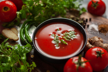 Close-up of Indian Homemade fresh and healthy tomato soup garnished with fresh coriander leaves and ingredients and herbs, served in a black bowl over the wooden top background.