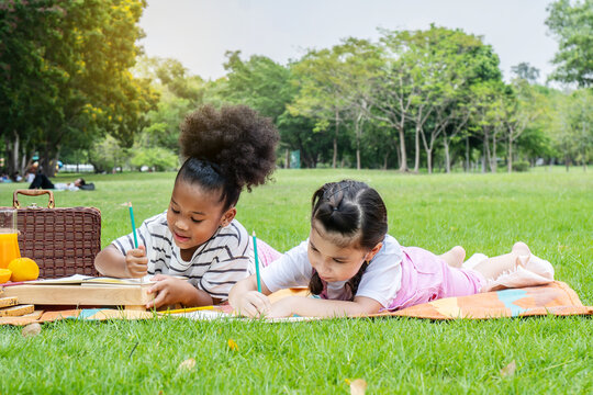 Two american african little girl lying and drawing with colored pencils at summer park, feel happiness, outdoor education concepts