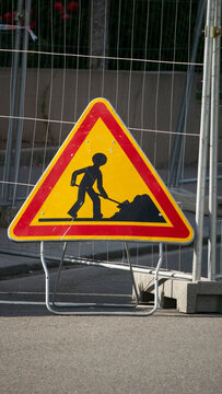 Yellow and red triangle road work sign next to a fence by a construction site representing a man shovelling gravel or dirt
