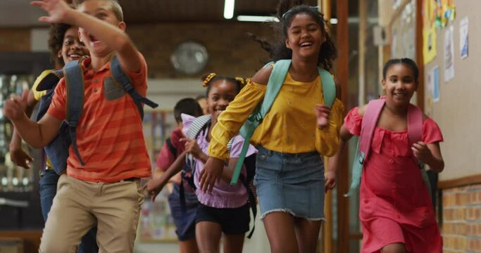 Diverse group of happy schoolchildren wearing backpacks, running fast through school corridor